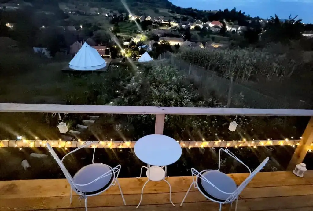 Wooden deck at dusk with wrought-iron chairs and string lights, overlooking a bell tent and the lights of the village below