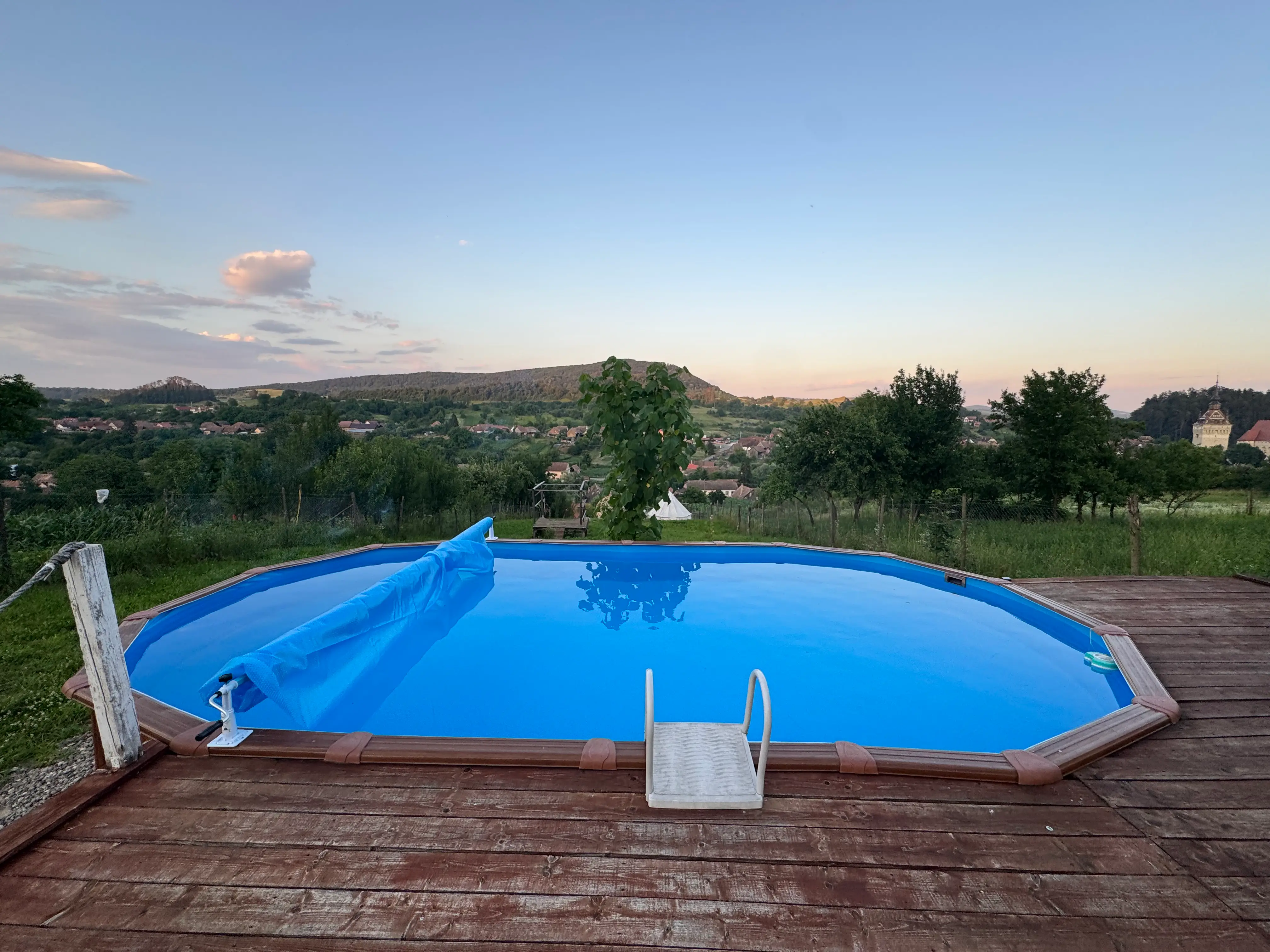 The pool on its wooden deck at dusk, the cover half rolled back, looking over the village rooftops to the fortified church tower and the hills beyond