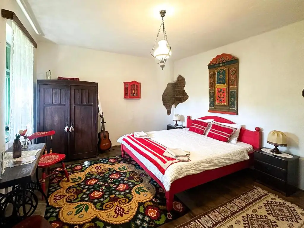 Bedroom with a red-painted wooden bed dressed in striped folk textiles, an antique painted Saxon cabinet on the wall and woven rugs on the floor